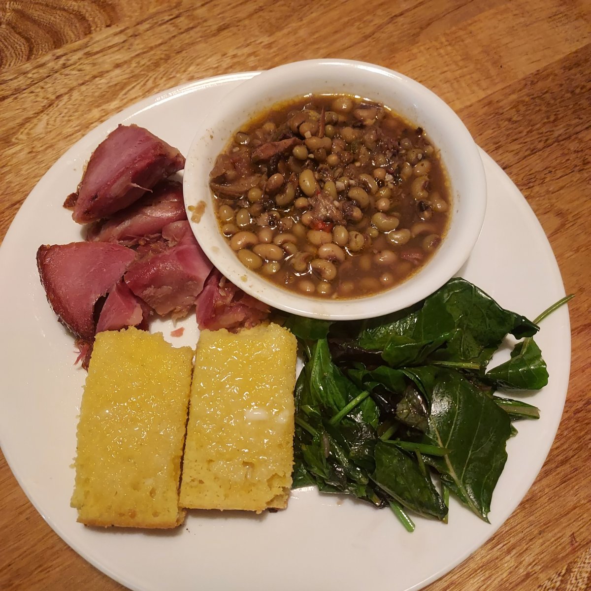 White plate on a wooden table. Three quadrants contain food directly on the plate, while one quadrant in the upper right corner contains a white bowl with cooked purple hull peas in smoked pork broth, a few small stray pork chunks scatter across the top of the peas. Moving clock-wise around the plate from the bowl are fresh greens, wilted and shiny with pork fat; a rectangular piece of cornbread split top from bottom and well-buttered; and finally, several chunks of roasted ham.